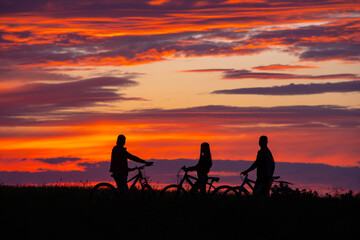 Fototapeta na wymiar Image of sporty company friends on bicycles outdoors against sunset mountain. Silhouette of three cyclist along sunset sky.