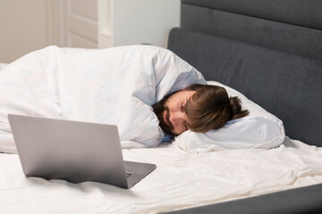 Close-up of a man of with a beard is sick wrapped in a white blanket lies in bed and uses a laptop. Concept: people using laptops mockup copy space 