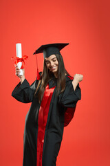 Front view of glad girl with bachelor, master degree holding diploma. Pretty female standing with closed eyes, showing hurray, smiling. Isolated on red studio background.