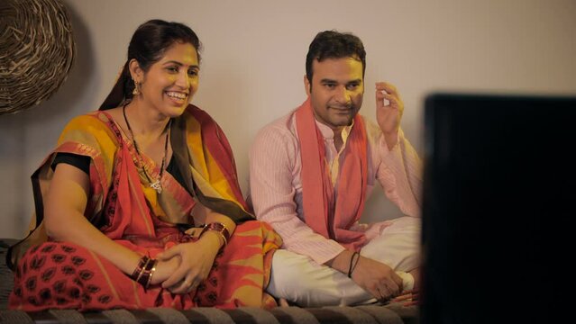 Indian village couple happily watching television together sitting on a woven cot. Medium shot of a joyous Indian husband-wife sitting together in their free time to watch their favorite TV program