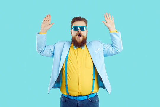 Funny fat young redhead man in stylish party suit and sunglasses pretends to be scared, opens his mouth and raises his hands up, studio shot on turquoise background