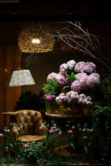 Florist shop window. Beautiful decoration of luxurious pink flowers, interior items, lighting. Showcase window with atmospheric lights, vintage armchair. Dark exquisite stage photo. Vertical image 