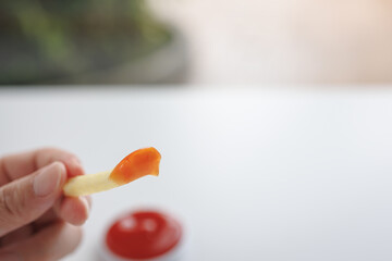 Man hand holding French fries with his fingers which dip ketchup (tomato sauce) on top of it isolated in blurred background. Fast food, unhealthy food concept.