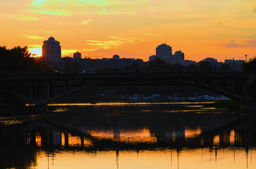Fototapeta premium Magic sunset in Kyiv. Silhouettes of buildings and a bridge reflected in tranquil water of Rusanivka's channel. Vibrant orange sunset colors