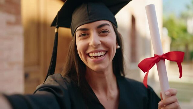 Young hispanic woman wearing graduated uniform holding diploma having video call at university