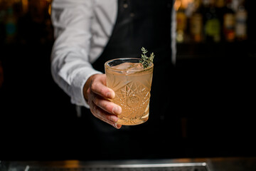 great view of glass with cocktail garnished with rosemary in the hand of bartender on blurred background