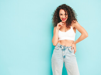 Sexy young beautiful smiling female in trendy summer hipster clothes. carefree woman posing near blue wall in studio. Positive brunette model with afro curls hairstyle. Cheerful and happy