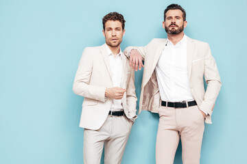 Portrait of two handsome confident stylish hipster lambersexual models. Sexy modern men dressed in white elegant suit. Fashion male posing in studio near blue wall