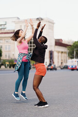 Smiling cheerful friends in sportswear running in the city discussing, giving hi five, celebrating success and happiness. Multiethnic women having a fitness workout.