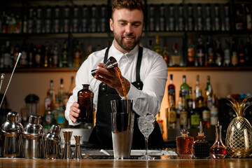 view on bar counter with various shakers and bottles on it and male bartender gently pours alcoholic drink into cup