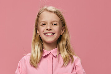 horizontal portrait of a school-age blonde girl in a pink shirt, standing against a one-time...