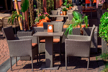 wicker rattan fittings on the terrace of a street cafe lit by sunlight, a square table with napkin holder and chairs with a soft cushion surrounded by ornamental plants flowers and palm trees, nobody.