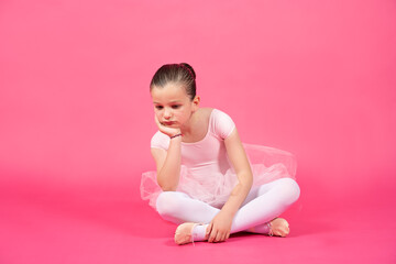 Bored little ballet dancer girl sitting on the floor. Studio shot with pink background