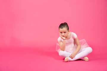 Bored little ballet dancer girl sitting on the floor and looking at camera. Studio shot with vivid pink background.