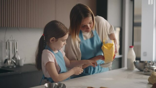 mother is teaching her little girl to make dough and bake bread in home, woman is pouring flour in sifter