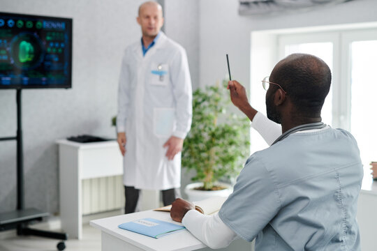 Rear View Of African Medical Student Raising His Hand And Asking Question To Speaker While Sitting At Desk At Seminar