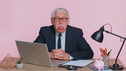 Senior mature older business man working on laptop, making funny face, fooling around, putting his tongue out remote working disrespecting someone at office. Elderly grandfather on pink background