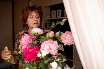 Active concentrated senior woman enjoy retirement, gardener hobby, taking care of peony flowers, watering, spraying. 