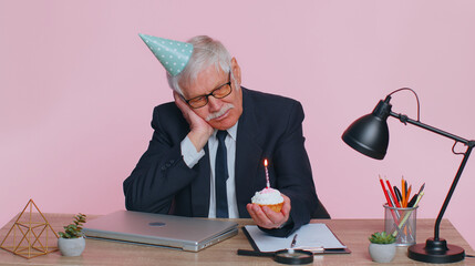 Sad upset lonely senior businessman in festive hat celebrating birthday party alone, feel frustrated despair disappointed sadness holds cake with candle at office. Elderly man isolated on pink studio