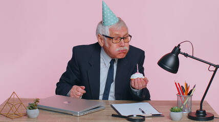 Happy senior mature businessman celebrating lonely birthday in office, blowing candle on small cake making a wish. Elderly success grandfather man wearing festive cap celebrate anniversary party alone