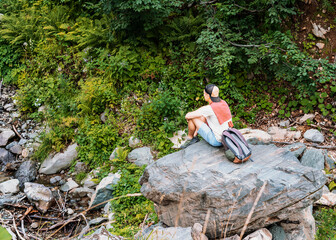 Fototapeta premium Young man in a cap with a backpack sitting on a large stone and looking at a mountain stream among the forest and lush vegetation in a summer hike Enjoying nature Active lifestyle