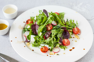 Green salad of fresh lettuce leaves, cherry tomatoes, feta cheese and assorted seeds with a sauce of honey, olive oil and balsamic vinegar on a flat plate on a stone table. Healthy vegan food.