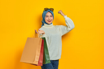 Cheerful beautiful Asian woman in white sweater holding shopping bags and celebrate luck isolated over yellow background