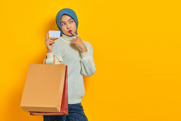 Pensive Beautiful Asian woman in white sweater holding shopping bag and credit card, thinking about something isolated over yellow background