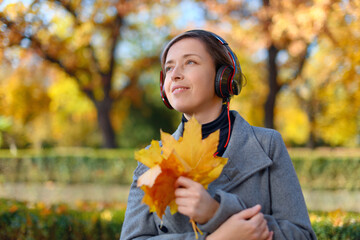 beautiful woman portrait, she is in autumn park and listens to music with headphones, trees with yellow leaves, bright sunny day