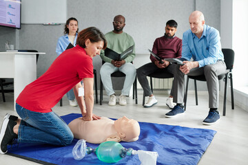 Medical instructor showing CPR on training mannequin on floor to people who sitting on chairs and...