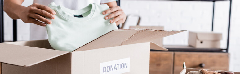 Cropped view of african american seller putting clothes in box with donation lettering, banner.