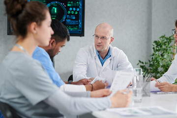 Obraz premium Mature male doctor in white coat sitting at table with his colleagues and discussing work during meeting at office