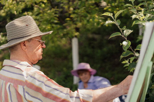 HAPPY SENIOR COUPLE Spend Time Together In Their Garden Or Nature. A Man Is Painting His Wife While She Is Relaxing With A Book. Growing Old Beautifully Together. Vibrant Green Garden