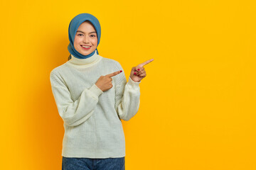 smiling beautiful Asian woman in white sweater pointing fingers aside at copy space and looking...