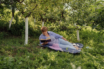 Senior woman smiling and reading a book while lying and relaxing on a blanket in the green garden