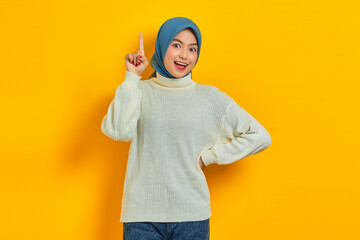 Cheerful young Asian woman in white sweater pointing finger up at copy space isolated over yellow background