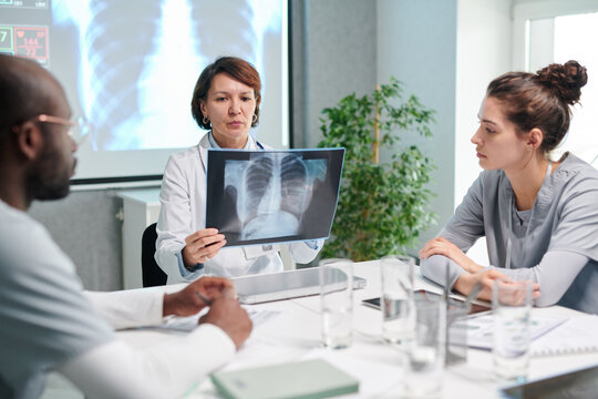 Mature female doctor in white coat examining x-ray image of lungs at table and discussing it with her colleagues during meeting