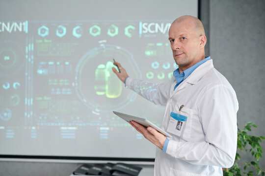 Portrait of mature male doctor in white coat with digital tablet pointing at projector with medical charts at conference