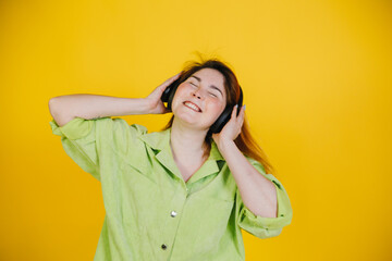 a red-haired beautiful woman stands on a yellow background in headphones listens to music and have fun and smiling