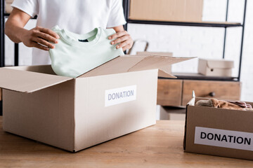 Cropped view of african american seller putting clothes in box with donation lettering on table.