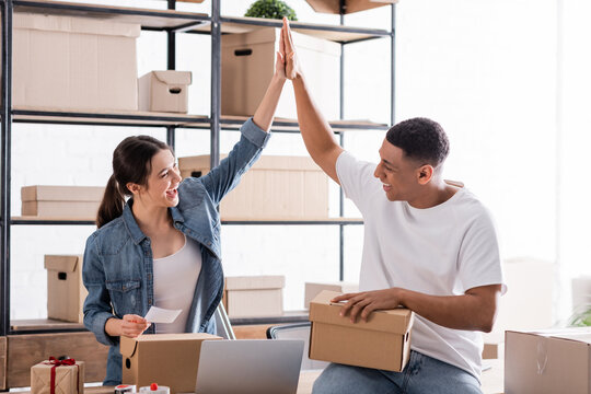 Cheerful multiethnic sellers giving high five near cardboard boxes and laptop in online web store.
