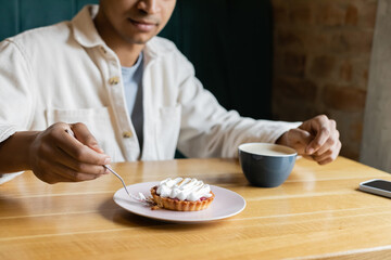 cropped view of young african american man holding fork near tasty tart on plate near cup and smartphone.