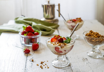 Yogurt with strawberry compote and granola with fresh fruits in dessert cups on white wooden background. Coffee pot backside