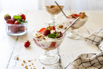 Yogurt with strawberry compote and granola with fresh fruits in dessert cups on white wooden background