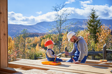 Father with toddler son building wooden frame house. Male builders hammering nail into plank on...