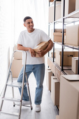 Positive african american seller holding carton box near rack and ladder in online web store.