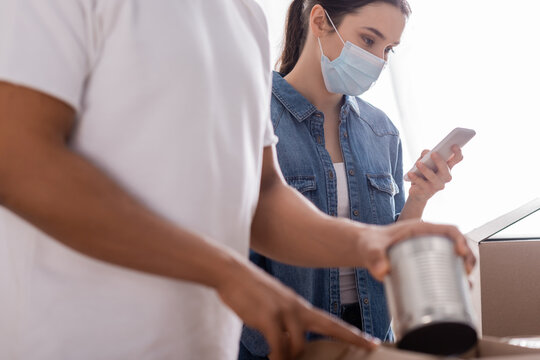 Seller in medical mask using smartphone near blurred african american colleague packing canned food in online web store.