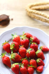 Lilac plate full of fresh strawberries, straw bag and retro sunglasses on wooden table. Selective focus.