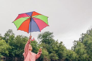 A little schoolgirl girl raises a multi-colored umbrella up to the sky in nature, she smiles and is happy, free from lessons