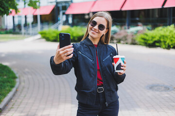 Beautiful and young girl in sunglasses writes a message on a cell phone and drinking coffee outdoors